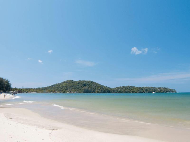 Weißer Sandstrand mit ruhigem türkisem Meer und bewaldetem Hügel unter klarem Himmel.