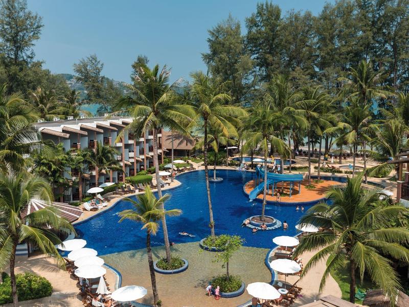 Großer tropischer Hotelpool mit Palmen, Liegestühlen und zweistöckigen Zimmern im Hintergrund.