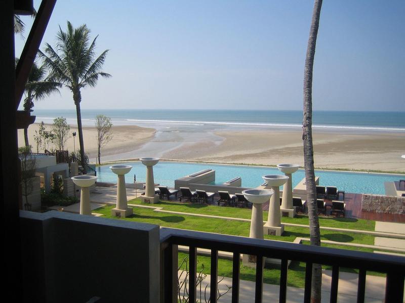 Strandblick mit Palmen, Sitzgelegenheiten und blauem Meer im Hintergrund unter klarem Himmel.