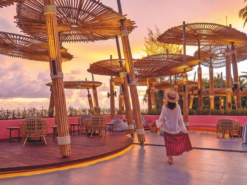 Frau in Sommerkleidung geht auf geschwungenem Weg mit großen Strohhut-Dächern bei Sonnenuntergang.