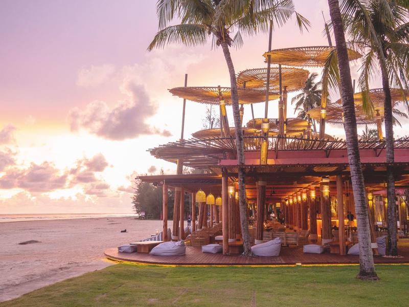 Strandresort mit Palmen und beleuchtetem Restaurant bei Sonnenuntergang am Meer.