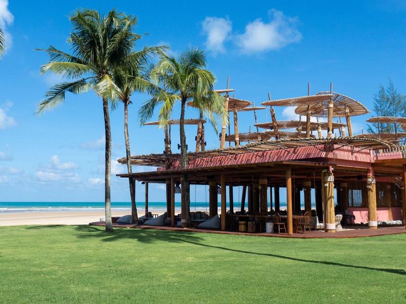 Strandresort mit Palmen, Holzterrasse und blauem Himmel, grüne Wiese im Vordergrund.