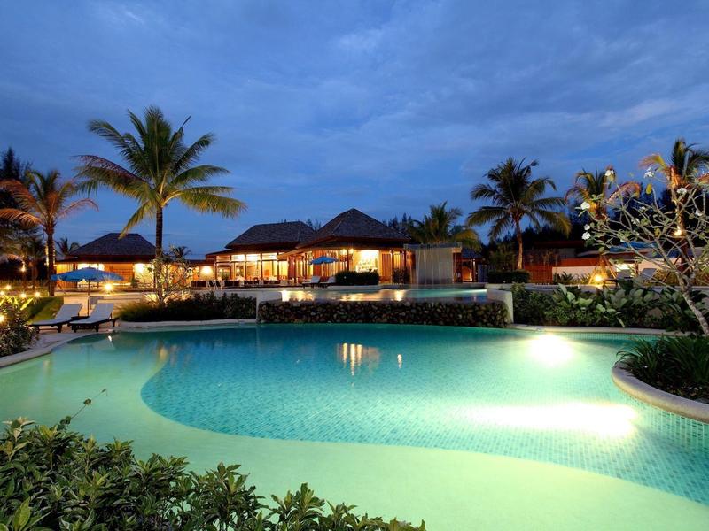Ein beleuchteter Pool umgeben von Palmen und Gebäuden bei Abenddämmerung mit blauem Himmel.