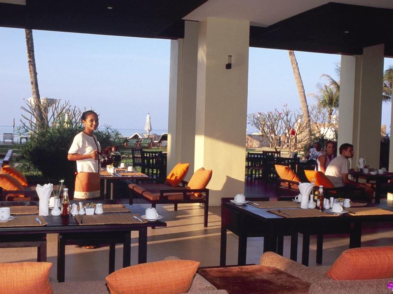 Offenes Restaurant mit Tischen, Stühlen und Bedienungen, Sonnenschein und Pflanzen im Hintergrund.