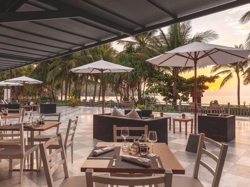 Freiluftrestaurant mit Tischen, Stühlen und Sonnenschirmen neben einem Strand mit Palmen und Sonnenuntergang.