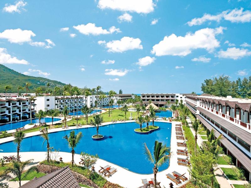 Große Hotelanlage mit Pool, Palmen und Bergen im Hintergrund unter blauem Himmel.