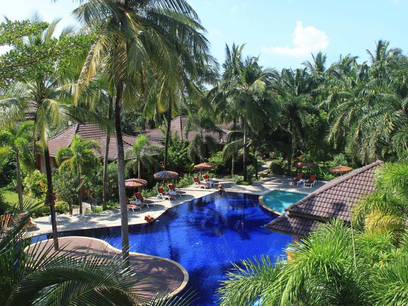 Großer blauer Pool umgeben von Palmen, grünen Pflanzen und Liegestühlen unter Sonnenschirmen.