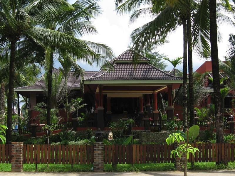 Tropisches Haus mit Holzdach umgeben von Palmen und grünem Garten hinter einem Zaun.
