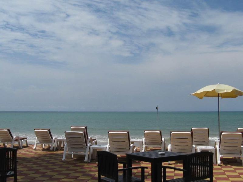 Strand mit Liegestühlen, Sonnenschirm, Palmen und ruhigem Meer unter bewölktem Himmel.