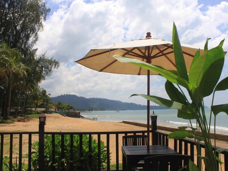 Strand mit Sand, blauem Meer und bewölktem Himmel, Tisch mit Sonnenschirm und Pflanzen im Vordergrund.
