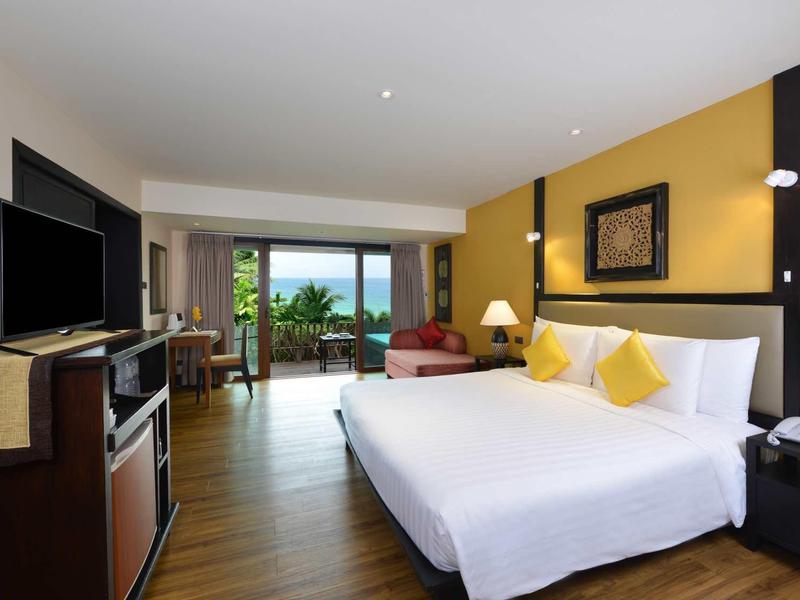 Modern hotel room with large bed, TV, seating area, and view of green landscape and sea.