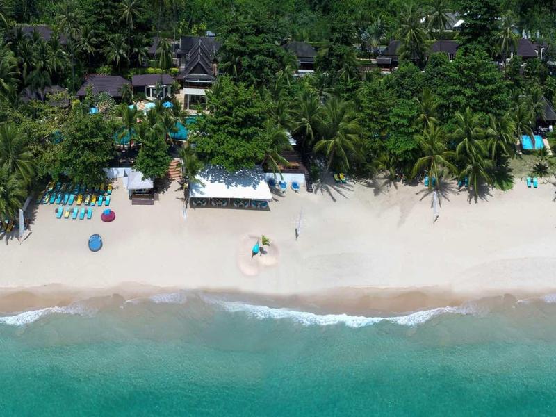 Aerial view of a tropical beach with white sand, turquoise sea, and palm-lined buildings.