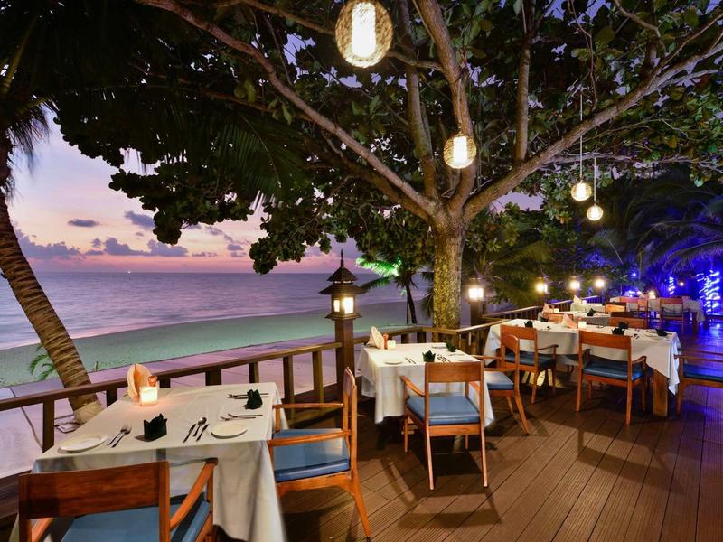 Beachfront restaurant with set tables under hanging lamps at sunset.