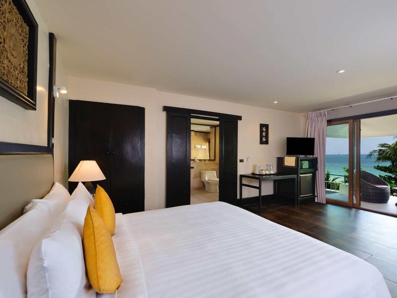 Modern hotel room with large bed, desk, and ocean view through sliding balcony door.