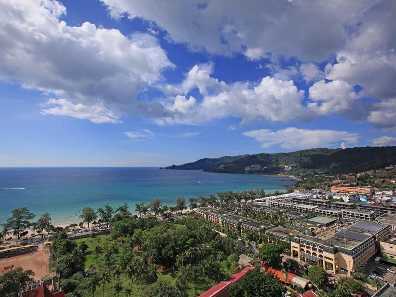Küste mit blauem Meer, Sandstrand, grünen Palmen und Gebäuden unter bewölktem Himmel.