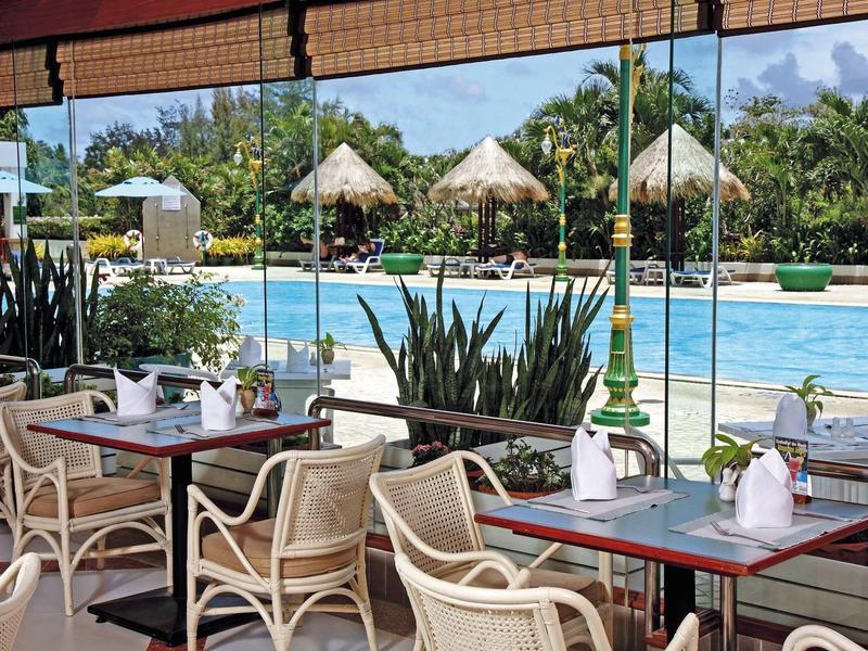Terrassenrestaurant mit Holzstühlen und Tischen vor großem Pool und grüner Palmenlandschaft.