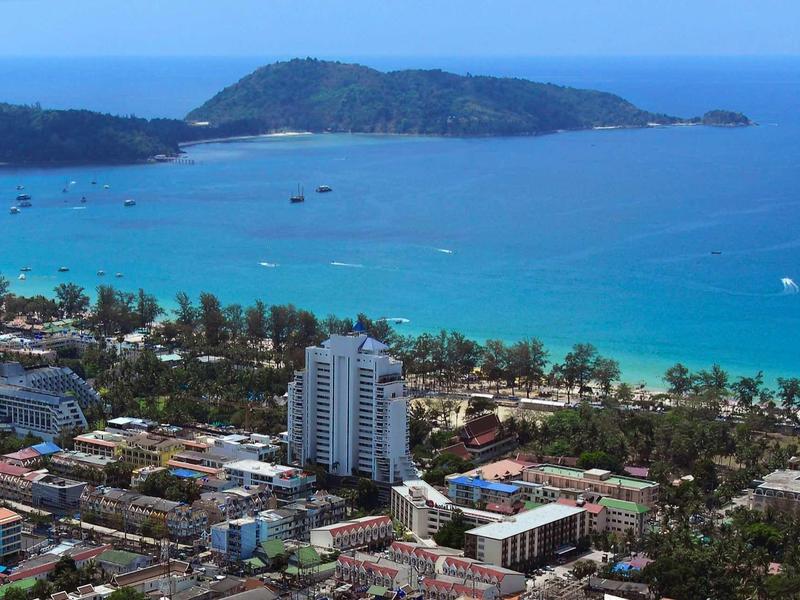 Küstenstadt mit hohen Gebäuden, grünen Bäumen und blauem Meer mit Insel im Hintergrund.