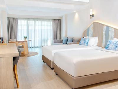 Helle Hotelzimmer mit zwei großen Betten, Holzmöbeln und großen Fenstern mit Vorhängen.