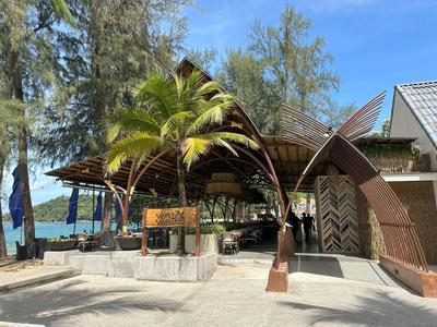 Offener Hotelbereich mit tropischem Schattendach, Palmen und Blick auf das Meer bei klarem Himmel.