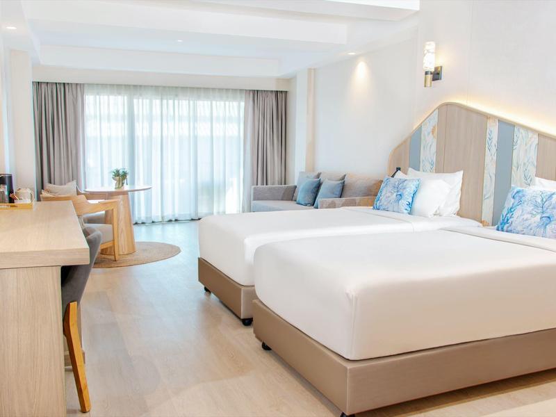 Helle Hotelzimmer mit zwei großen Betten, Holzmöbeln und großen Fenstern mit Vorhängen.