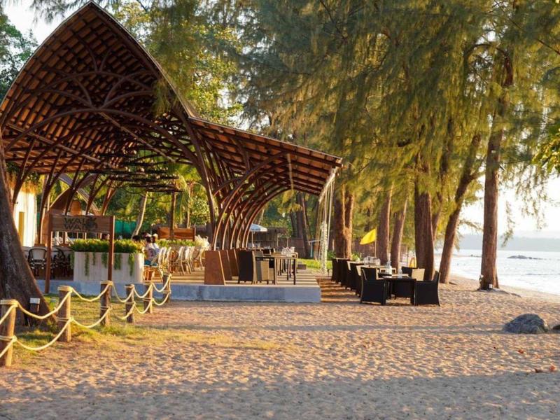 Holzpergola am Strand mit Stühlen, Sandboden, Bäumen und Blick auf das Meer bei Sonnenlicht.