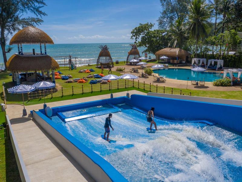 Zwei Personen surfen in einem Pool mit künstlichen Wellen in einer tropischen Hotelanlage am Meer.