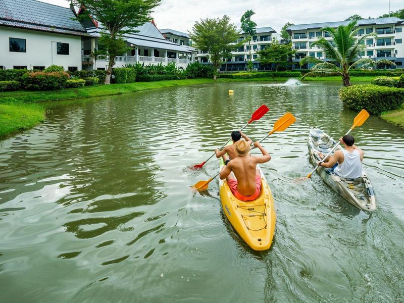 Zwei Männer paddeln auf Kajaks in einem grünen, ruhigen Fluss neben Wohnhäusern.