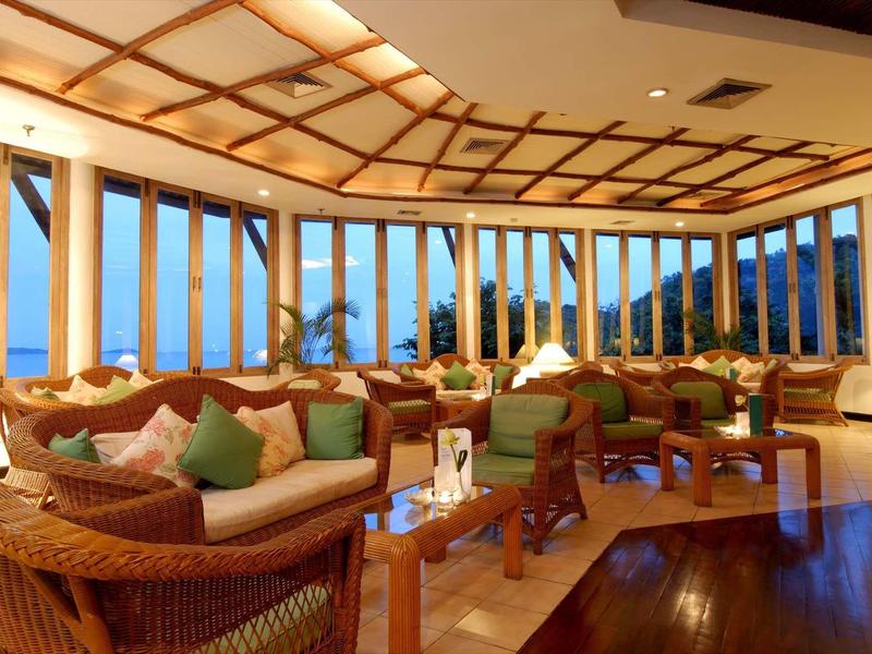 Großer heller Raum mit großen Fenstern, Holzdecke, Korbmöbeln und grünen Kissen.