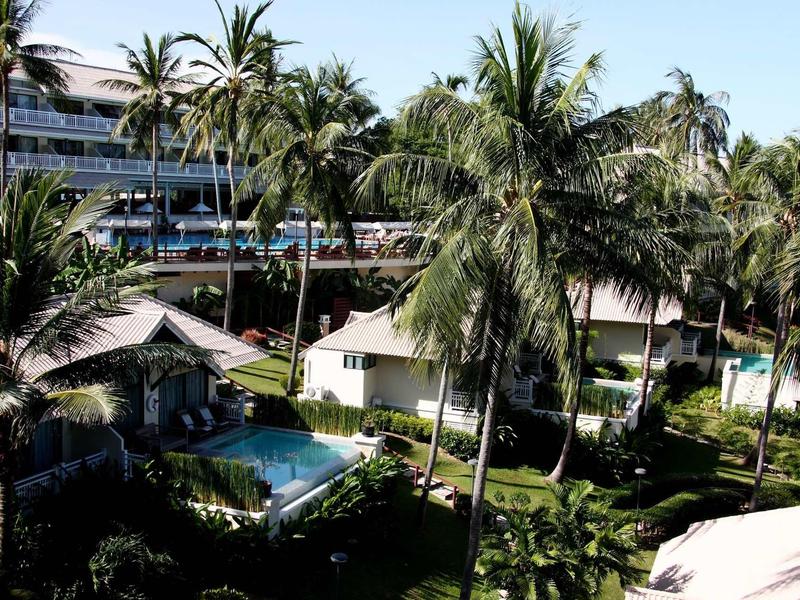 Grüner Hotelgarten mit Palmen, kleinen Hütten und blauem Pool vor mehrstöckigem Gebäude.