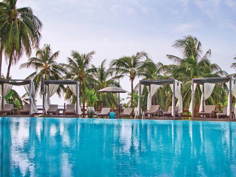 Großer Pool mit klarem Wasser, umgeben von weißen Liegen, Pavillons und hohen Palmen bei Himmel.