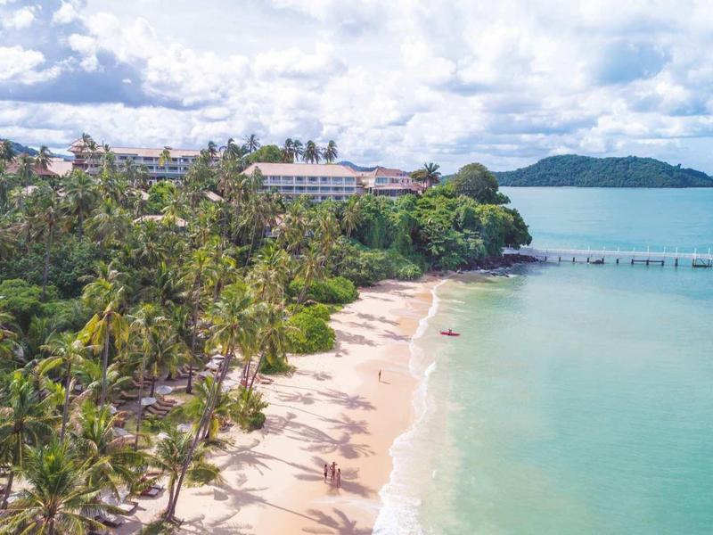 Strand mit Palmen und Gebäuden an der Küste, türkisblaues Meer und bewölkter Himmel.