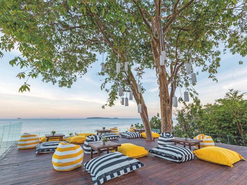 Outdoor-Sitzbereich mit bunten Kissen auf Holzboden, großer Baum und Blick aufs Meer bei Sonnenuntergang.