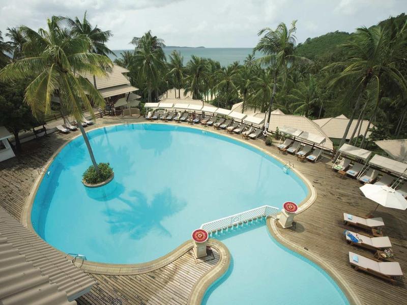 Ein blauer Pool mit Liegestühlen und Palmen am Meer im Hintergrund unter bewölktem Himmel.