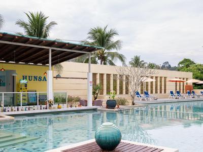 Hotelpool mit Sitzgelegenheiten, Sonnenschirmen und Palmen unter bewölktem Himmel.