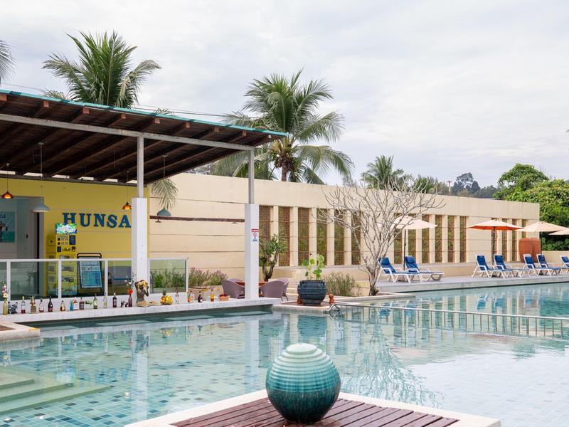 Hotelpool mit Sitzgelegenheiten, Sonnenschirmen und Palmen unter bewölktem Himmel.