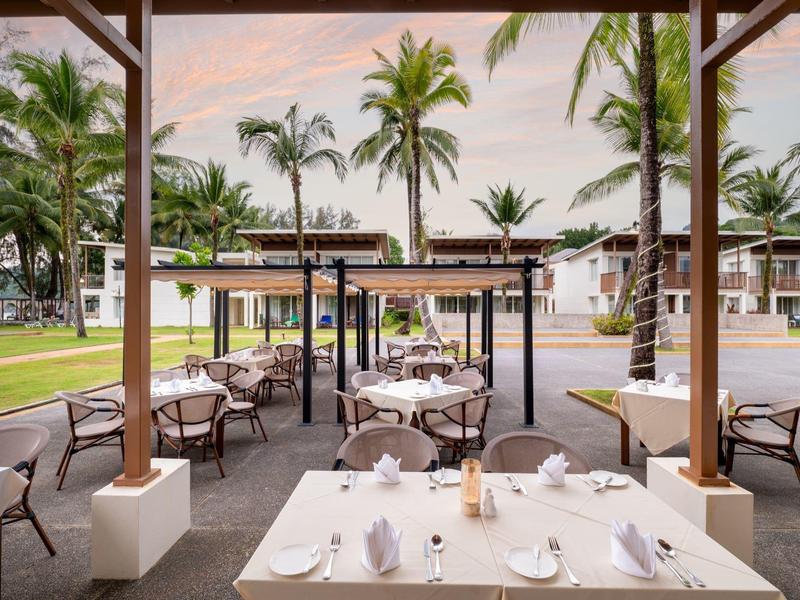 Outdoor-Restaurantbereich mit gedeckten Tischen und Blick auf Palmen und Hotelgebäude.