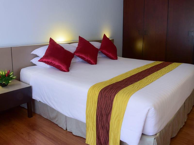 Habitación de hotel con cama grande, almohadas rojas, manta amarilla y muebles oscuros.