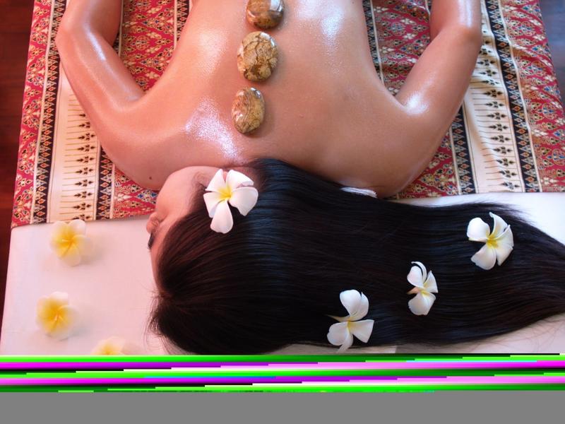 Vue de dos d'une personne recevant un massage aux pierres chaudes avec des fleurs dans les cheveux, sur un tapis à motifs.