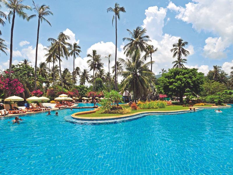 Grand piscine d'hôtel avec palmiers, chaises longues et parasols sous un ciel partiellement nuageux.