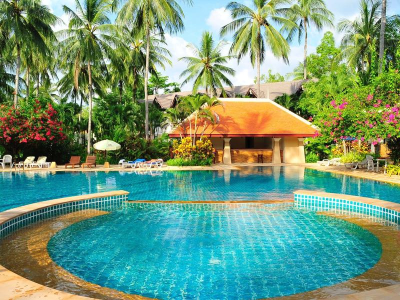 Piscine ronde entourée de chaises longues et de palmiers devant un bâtiment au toit orange.