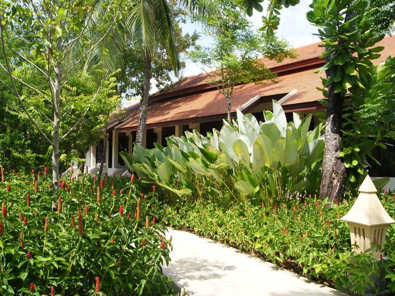 Sentier bordé de plantes tropicales menant à un bâtiment au toit rouge dans un environnement hôtelier verdoyant.