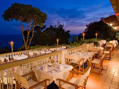 Terrassenrestaurant bei Dämmerung mit weißen Tischen, Holzstühlen und Blick auf Bäume und Himmel.