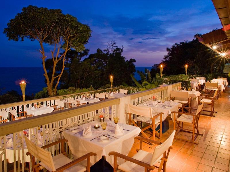 Terrassenrestaurant bei Dämmerung mit weißen Tischen, Holzstühlen und Blick auf Bäume und Himmel.