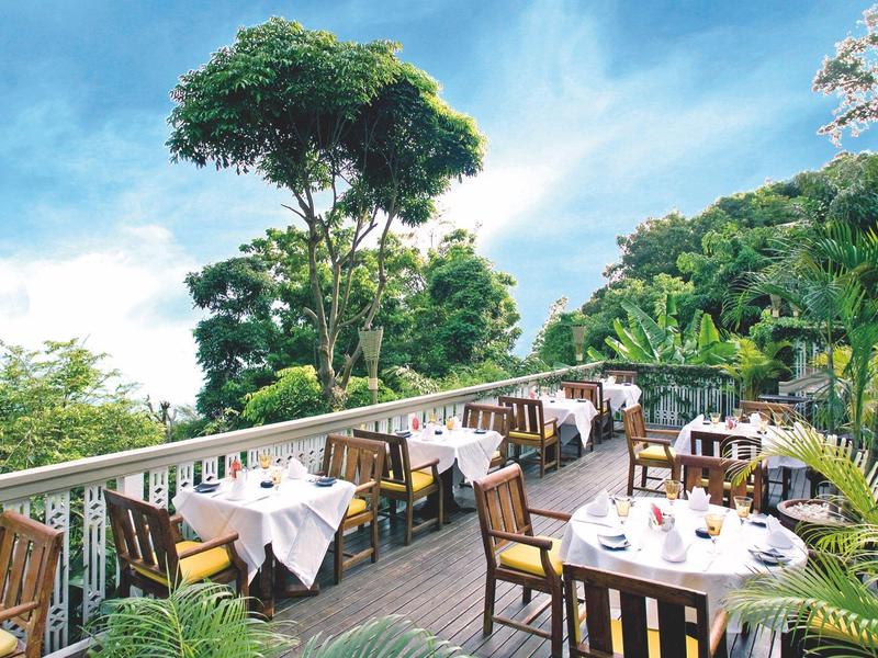 Freiluftrestaurant mit Holztischen und weißen Tischdecken, umgeben von grünen Bäumen und blauem Himmel.