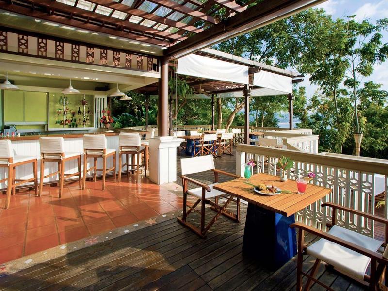 Terrasse mit Holzstühlen, Tisch, Barhockern und offener Bar, umgeben von Grün in hellem Licht.