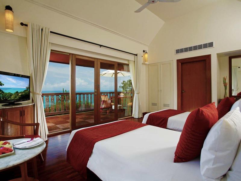 Hotelzimmer mit zwei Betten, rot-weißer Bettwäsche, Balkon, Meerblick und hellem Holzmobiliar.