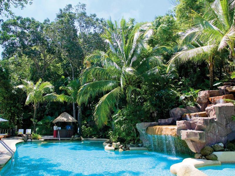 Ein blauer Pool mit Wasserfall umgeben von grünen Palmen und Bäumen unter blauem Himmel.