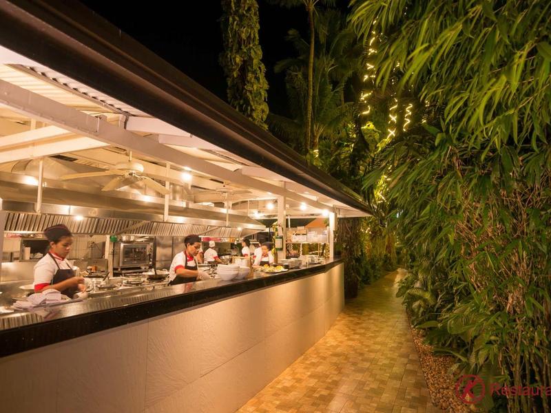 Eine lange Außenküche nachts, beleuchtet, mit mehreren Köchen und einem Weg neben Bambuspflanzen.