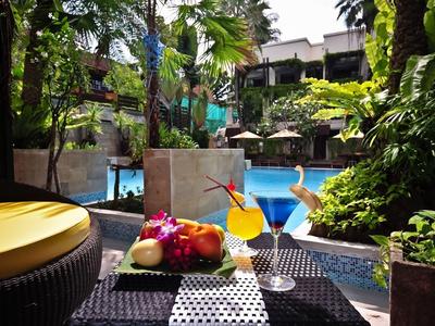 Bunte Früchte und Cocktails auf Tisch am Pool mit grünen Pflanzen und Sonnenschirmen im Hintergrund.