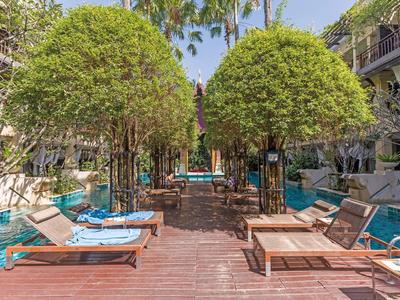 Poolbereich mit Holzsteg, Liegen und symmetrisch geschnittenen Bäumen unter blauem Himmel.